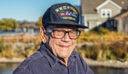 Smiling World War II veteran wearing a Navy cap standing outdoors near his home — representing veterans eligible for VA Aid and Attendance benefits.