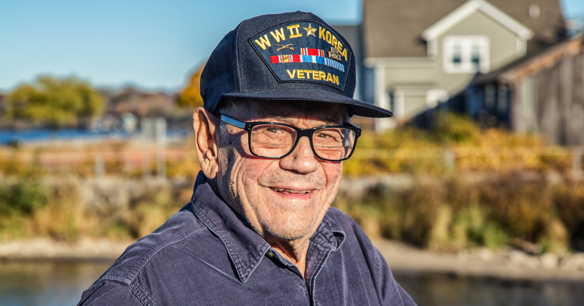 Smiling World War II veteran wearing a Navy cap standing outdoors near his home — representing veterans eligible for VA Aid and Attendance benefits.